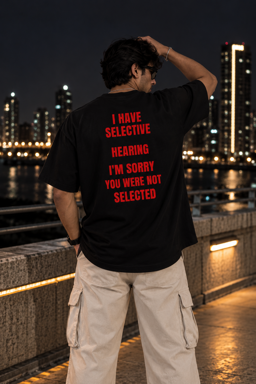 A man stands on a city bridge at night, viewed from behind, wearing a black t-shirt with bold red text that reads 'I HAVE SELECTIVE HEARING I'M SORRY YOU WERE NOT SELECTED' and light beige cargo pants.