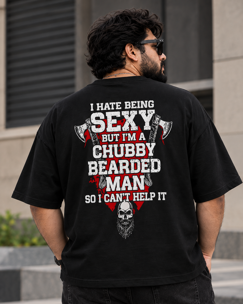 A man wearing a black graphic t-shirt from the back, standing outdoors in front of a modern building. The shirt features a bold white and red text design that reads 'I HATE BEING SEXY BUT I'M A CHUBBY BEARDED MAN SO I CAN'T HELP IT' with illustrations of an axe, a skull, and a large beard.