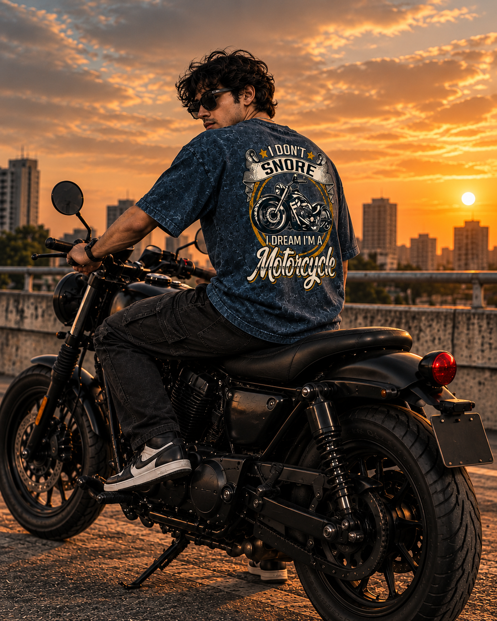 A man wearing a blue graphic t-shirt and black pants sits on a black cruiser motorcycle on an outdoor bridge at sunset. The back of the t-shirt features a large graphic print with the text 'I DON'T SNORE I DREAM I'M A Motorcycle'.